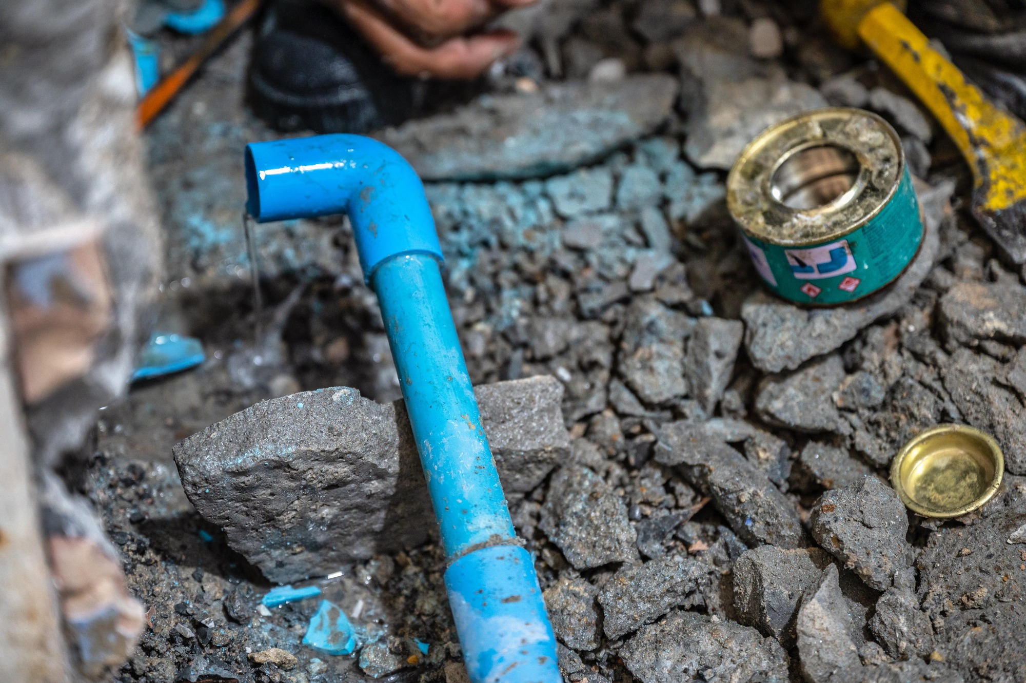 A broken blue PVC pipe with water leaking from it is surrounded by scattered pieces of debris and rocks. Nearby, there are a small green container and a yellow tool, possibly indicating ongoing repair work. The workspace is cluttered and appears to be in the process of being fixed.